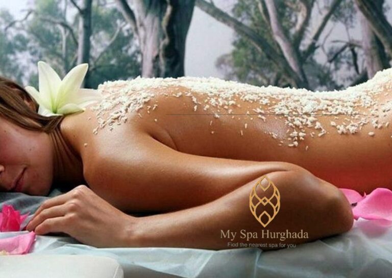 A close-up view focusing on a woman's back receiving a textured body exfoliation with small granular substances, simulating a scene from a professional Cleopatra massage program. She has an exotic flower tucked into her hair, and her expression is relaxed and serene. The texture of the scrub and the massage action are visible.